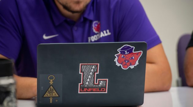 Laptop with multiple Linfield stickers on top opened on desk with student behind it.