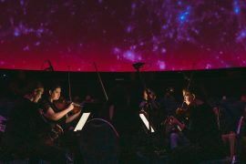 Four members of Third Angle play stringed instruments inside OMSI's planetarium with a glowing red sky above them.