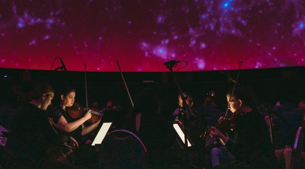 Four members of Third Angle play stringed instruments inside OMSI's planetarium with a glowing red sky above them.