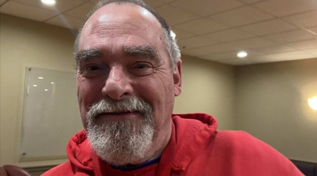An older man wears a red sweatshirt with faded letters that say "Linfield" and smiles slightly at the calendar.