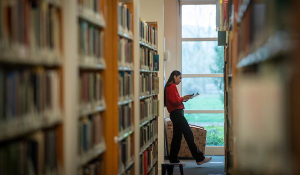 A student reads a book at the end of a library shelf.
