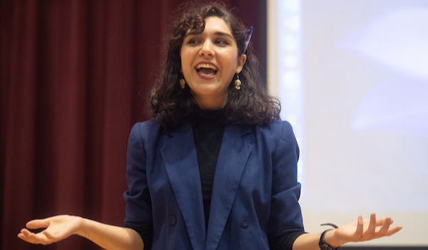 A student presents to a large room, wearing a blazer with a pen tucked behind her ear.