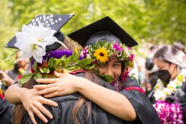 Two students embrace during commencement; their caps are circled with floral wreaths.