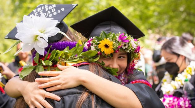 Two students embrace during Commencement; their caps are circled with floral wreaths.