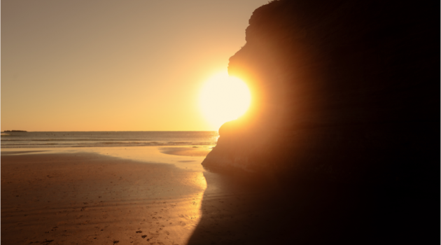 A photograph of a cliff at the Oregon coast; the sun is right at the edge of the cliff and its light breaks into radiating half-circles
