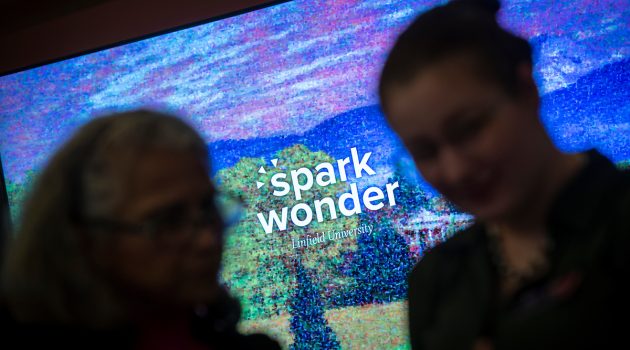 Two people stand in front of a projection of the Spark Wonder graphic, which renders Linfield's campus in Impressionist-style painting