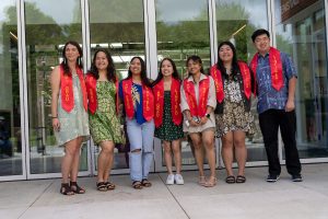 A group of students pose outside Keck Hall during Linfield University’s Asian American-Pacific Islander celebration; the students all wears red stoles that says “API Grad”.