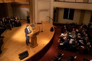 A man stands at a podium in Ice Auditorium and addresses the crowd during Linfield University’s Baccalaureate.