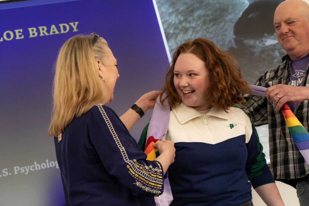 Family members place a lavender stole with rainbow trim at the bottom around the neck of a graduate during the Linfield University Lavender Celebration.
