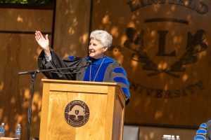 President Becky Johnson speaks during the 2024 Linfield University Commencement; they are standing at a podium with a large wooden Linfield University crest backdrop.