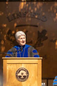 President Becky Johnson speaks during the 2024 Linfield University Commencement; they are standing at a podium with a large wooden Linfield University crest backdrop.