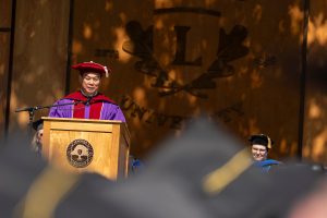 Shawn Chen speaks during the 2024 Linfield University Commencement; they are standing at a podium with a large wooden Linfield University crest backdrop.
