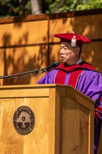 Shawn Chen speaks during the 2024 Linfield University Commencement; they are standing at a podium with a large wooden Linfield University crest backdrop.