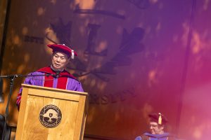 Shawn Chen speaks during the 2024 Linfield University Commencement; they are standing at a podium with a large wooden Linfield University crest backdrop.