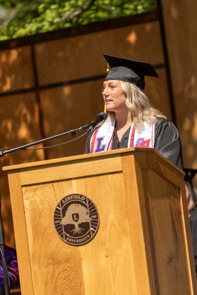 A graduate speaks during the 2024 Linfield University Commencement; they are standing at a podium with a large wooden Linfield University crest backdrop.