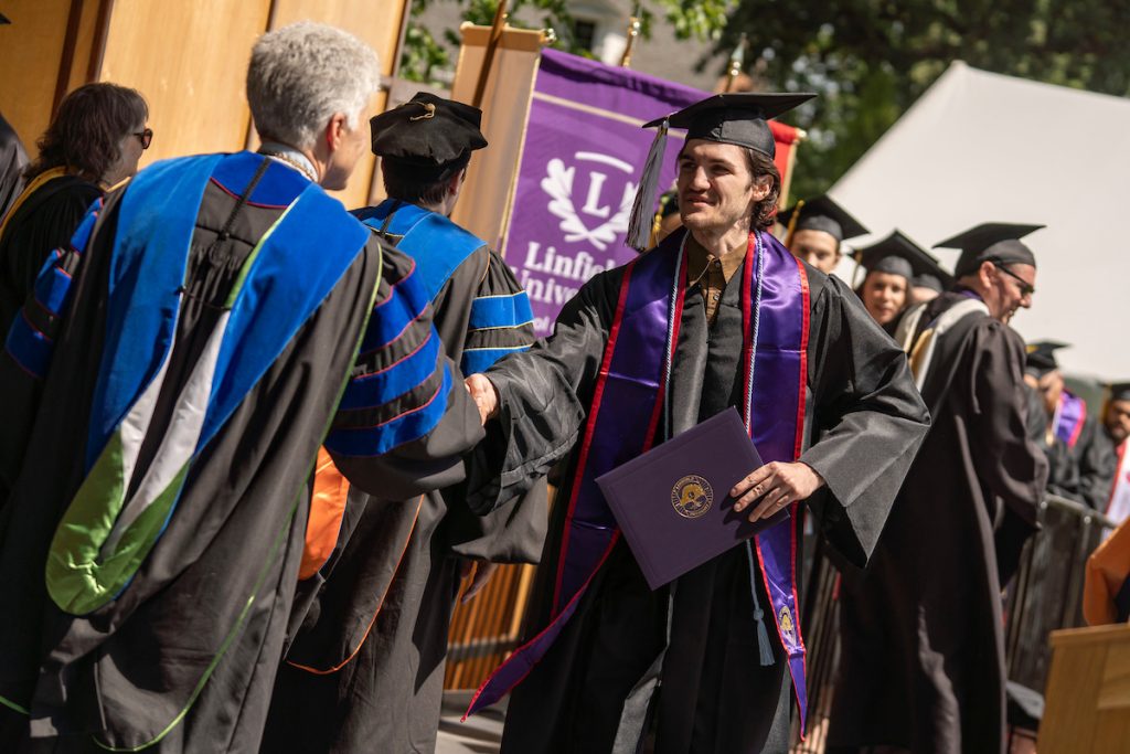 A student holds a diploma and shakes hands with Linfield University’s president during the 2024 Linfield University Commencement.
