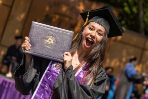 A student poses with their diploma and smiles during the 2024 Linfield University Commencement.