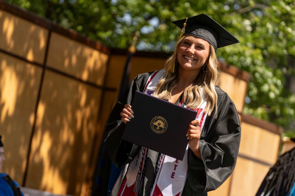 A student poses with their diploma and smiles during the 2024 Linfield University Commencement.