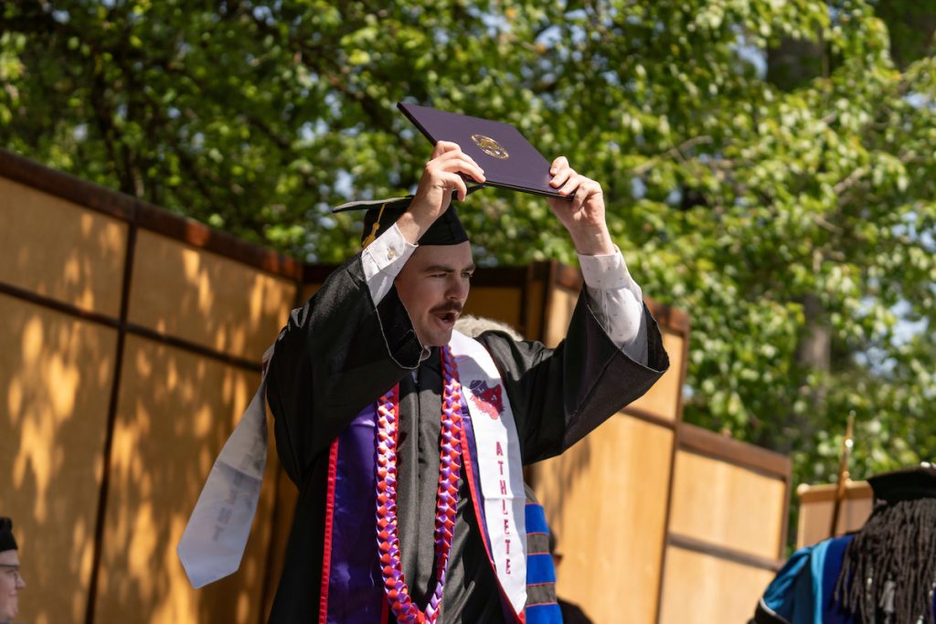 A graduate in his cap and gown holds his diploma above his head, smiling.