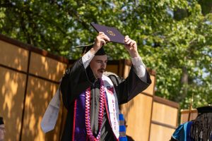 A graduate in his cap and gown holds his diploma above his head, smiling.