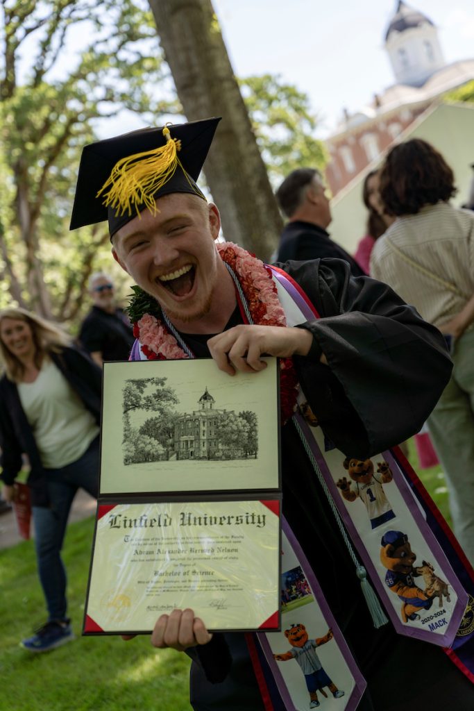A student poses with their diploma and smiles during the 2024 Linfield University Commencement.