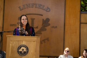 A speaker at the Nurses Pinning ceremony stands at a podium, with a ‘Linfield University’ wooden backdrop behind them.