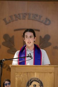 A speaker at the Nurses Pinning ceremony stands at a podium, with a ‘Linfield University’ wooden backdrop behind them.