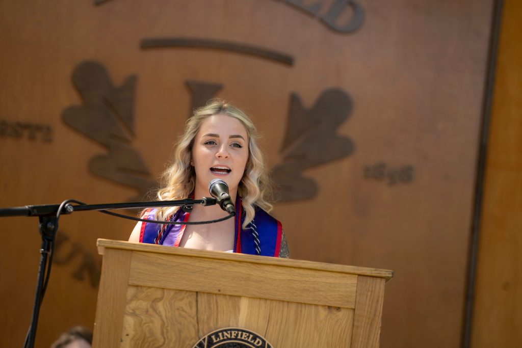 A speaker at the Nurses Pinning ceremony stands at a podium, with a ‘Linfield University’ wooden backdrop behind them.
