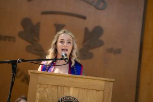 A speaker at the Nurses Pinning ceremony stands at a podium, with a ‘Linfield University’ wooden backdrop behind them.