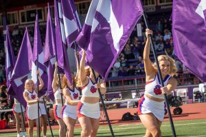 Cheerleaders hold Linfield flags as they run onto a football field. 