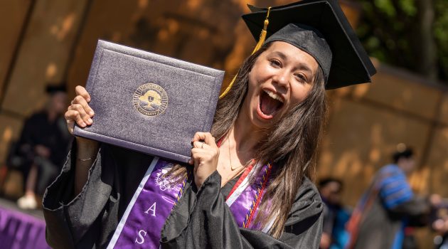 A student holds their Linfield diploma and smiles