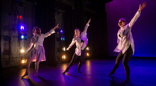 Three dancers extend into an elongated pose on a lit stage.