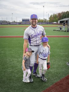 Two people pose in baseball uniforms with a little girl during Linfield Homecoming 2024