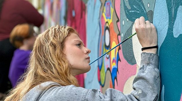 Student paints details on a large-scale mural.