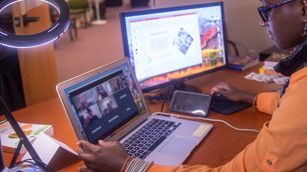 woman sits at a desk with laptop on a Zoom and second screen on a presentation