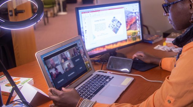 woman sits at a desk with laptop on a Zoom and second screen on a presentation