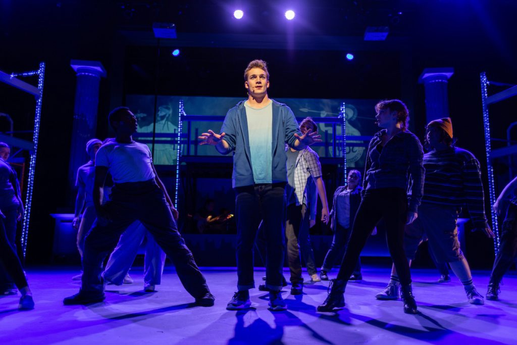 Scene from The Lightning Thief. Percy Jackson is lit with spotlight in center. Other characters surround him in the dark.