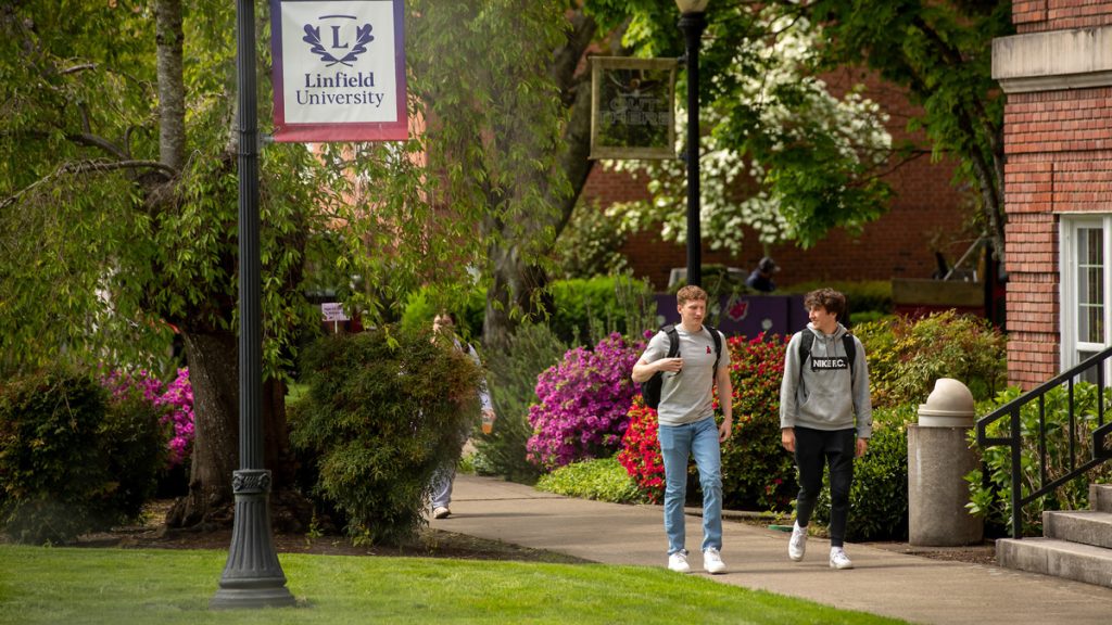 A pair of students walk on campus, talking, with flowers blooming around sidewalk and Linfield banner in foreground.