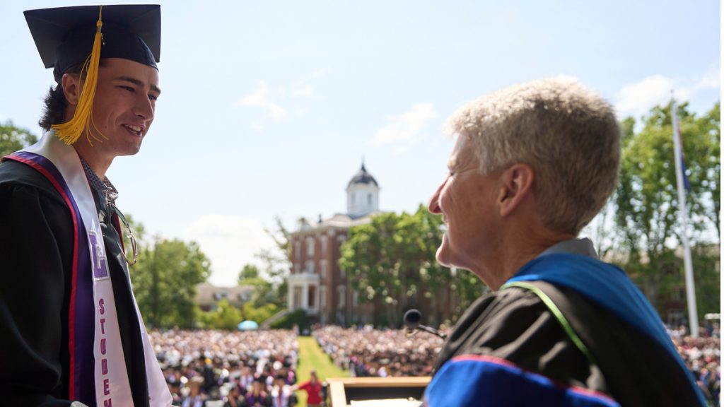 Interim President Becky Johnson shakes the hand of a graduate while thousands look on in the audience