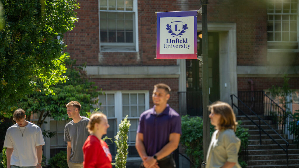 Students in foreground and background in front of building on McMinnville campus. Linfield University pole banner on center.