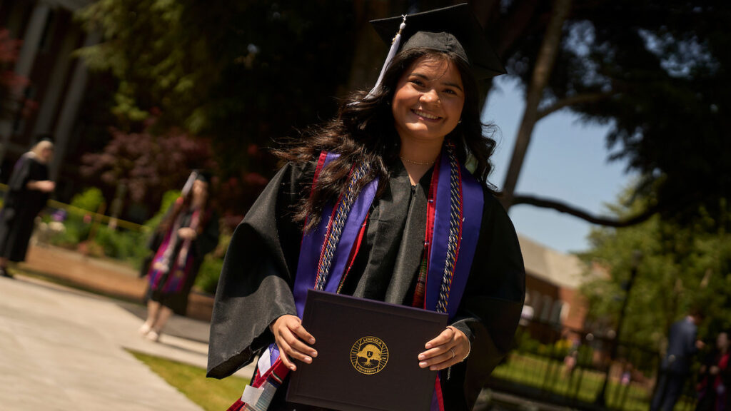 Linfield graduate in cap and gown smiles while holding her diploma