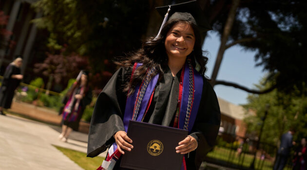 Linfield graduate in cap and gown smiles while holding her diploma