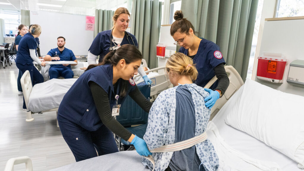 Nursing students help a standardized patient in the foreground stand with a strap. In the background, other students assist a patient sitting on a bed.