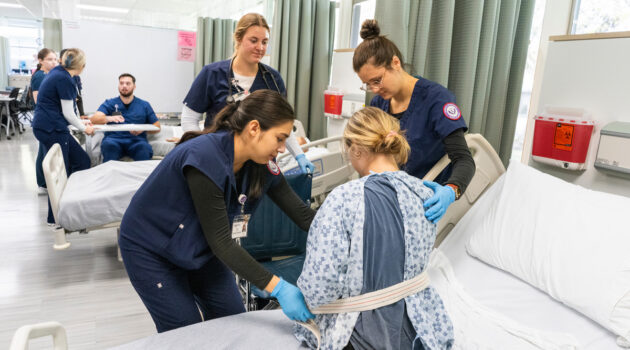 Nursing students help a standardized patient in the foreground stand with a strap. In the background, other students assist a patient sitting on a bed.