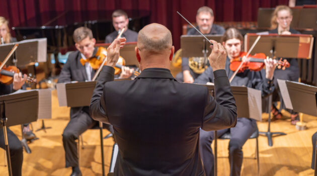 A composer directs members of an instrumental ensemble (seen with violins and brass)