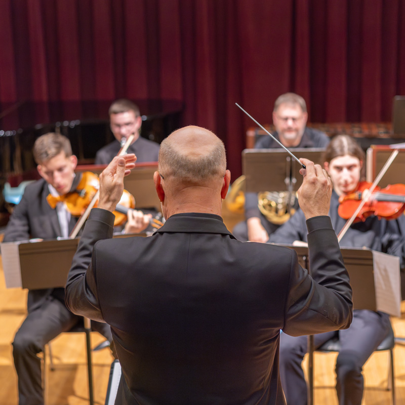 A composer directs members of an instrumental ensemble (seen with violins and brass)