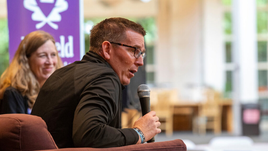 Mark Blegen speaks into a microphone at a meet-and-greet event, with Debbie Harmon Ferry looking on.