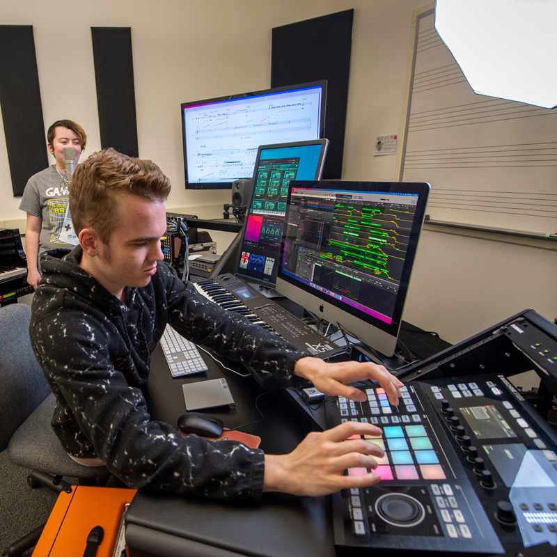 Two students work in composition studio. One in foreground works with computers and a soundboard. The other stands behind a microphone.