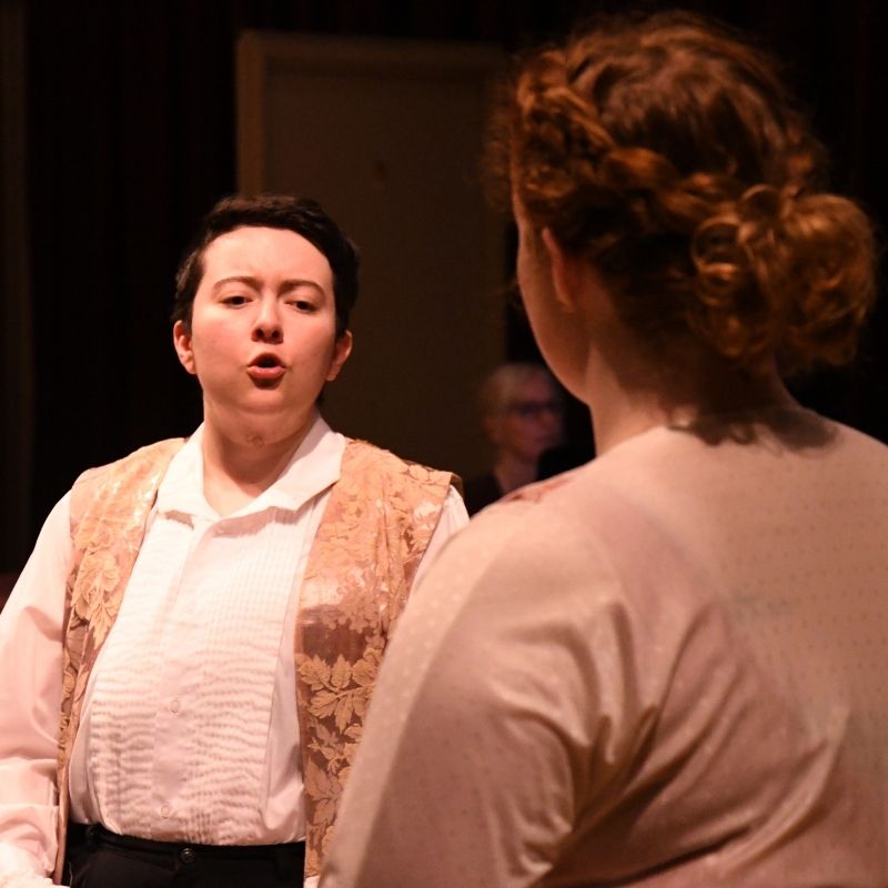 Two students perform an opera, singing to each other. One facing camera, one facing rear.
