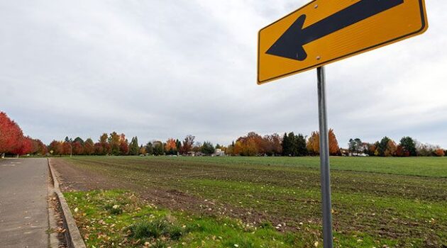 Yellow directional sign with arrow in foreground in front of large empty agricultural field.